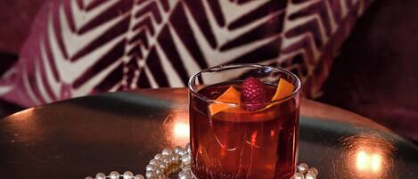 A glass of amber-colored drink with fruit garnish on a reflective surface, accompanied by a string of pearls, set against a patterned cushion.