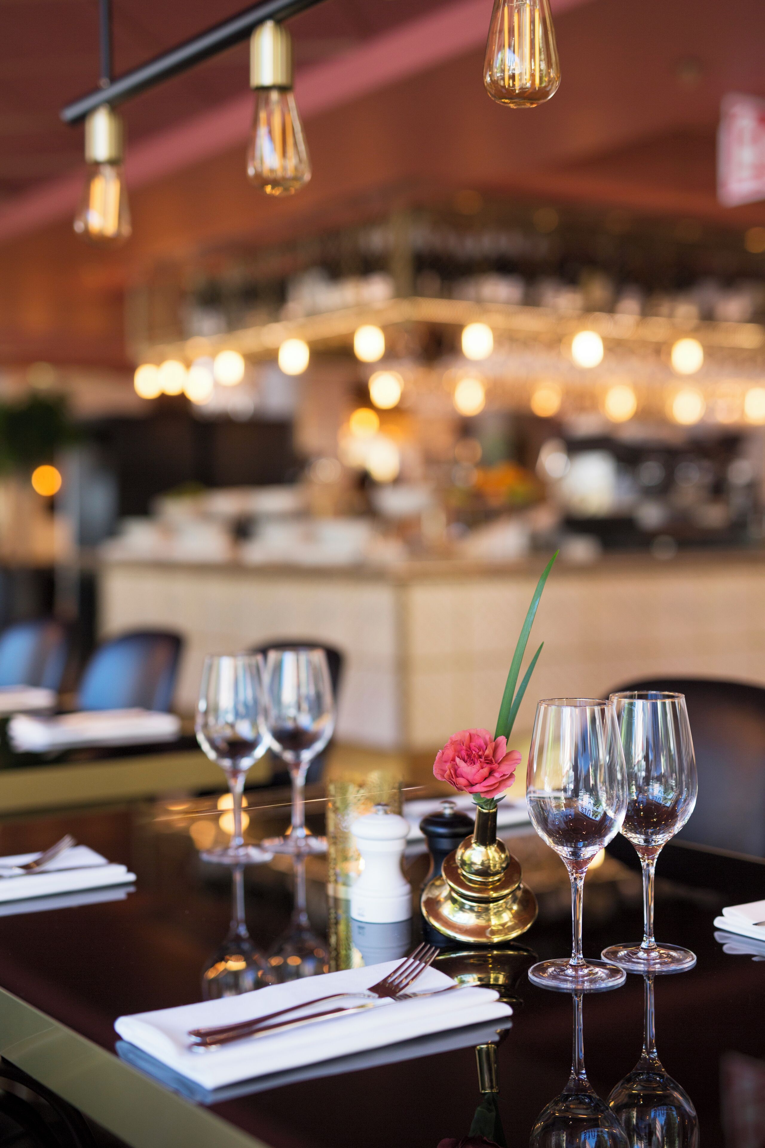 Un ambiente de restaurante con una mesa lista para comer, con copas de vino, un florero con una flor y una vela.