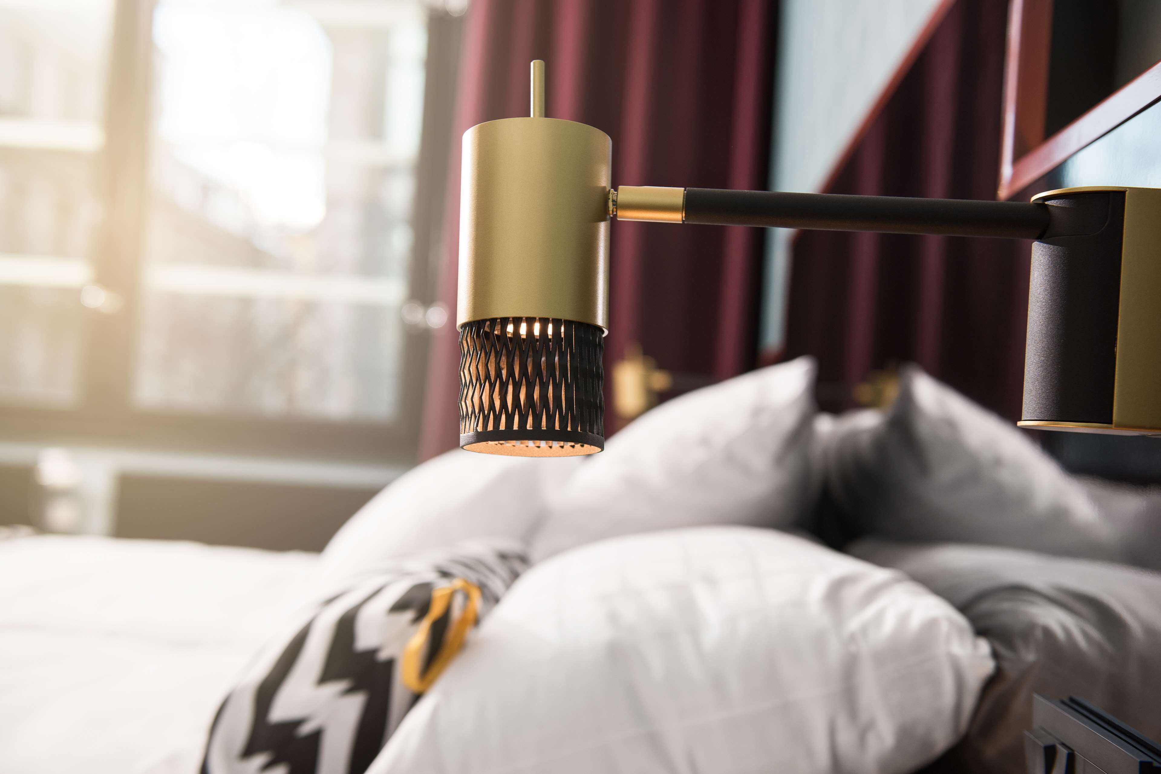 Una cama con almohadas blancas y estampadas, una lámpara dorada y negra, y una ventana con cortinas al fondo.