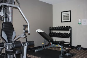 Un gimnasio con máquinas de pesas, una banca y un estante con mancuernas.