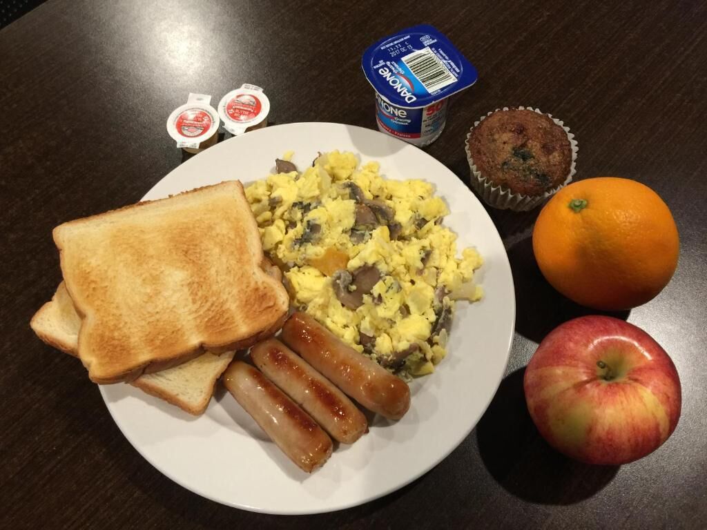 Um prato com ovos mexidos, salsichas, torradas, uma maçã, uma laranja e um muffin sobre uma mesa de madeira escura.