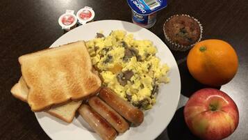 Um prato com ovos mexidos, salsichas, torradas, uma maçã, uma laranja e um muffin sobre uma mesa de madeira escura.
