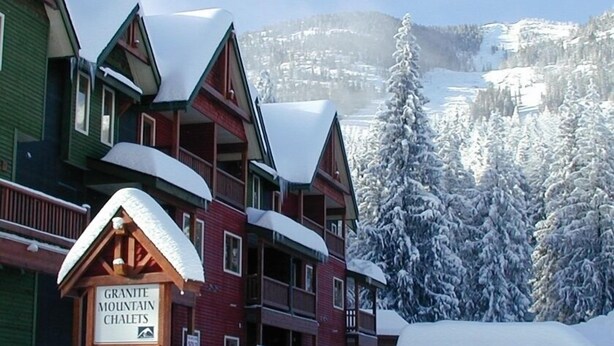A multi-story lodge with a sign for Granite Mountain Chalets, surrounded by snow-covered trees and mountains. - Red Mountain Village Condos (Rossland)
