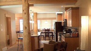 A spacious living room with a large wooden beam, a dining area with a table and chairs, and a kitchen with wooden cabinets and appliances. - Red Mountain Village Condos (Rossland)