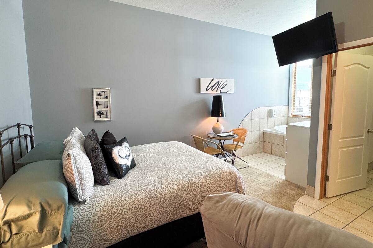 Une chambre à coucher avec un lit, une table de chevet avec une lampe, une petite table avec des chaises, un cadre photo et une porte donnant sur une autre pièce.