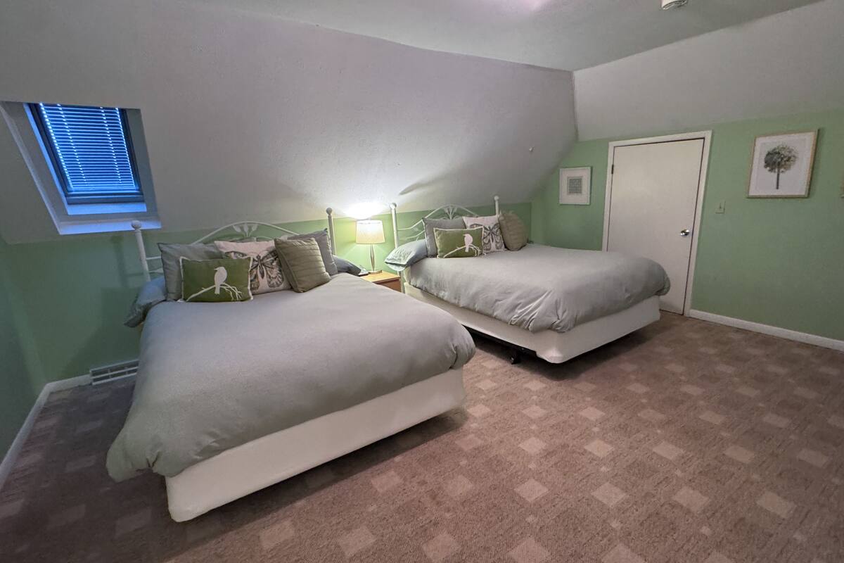Une chambre avec deux lits, une fenêtre avec des stores, une porte et un tableau accroché au mur.