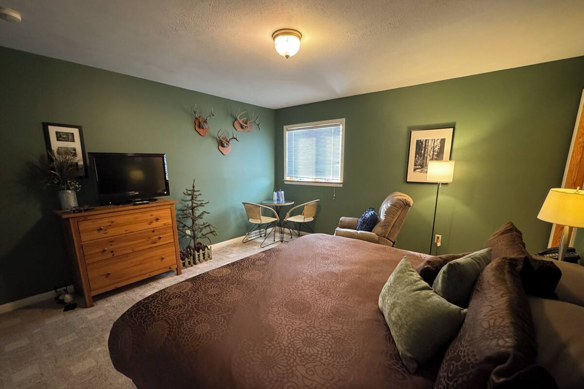 Une chambre à coucher avec un grand lit, une commode, un téléviseur, une petite table avec des chaises et une fenêtre avec des stores.