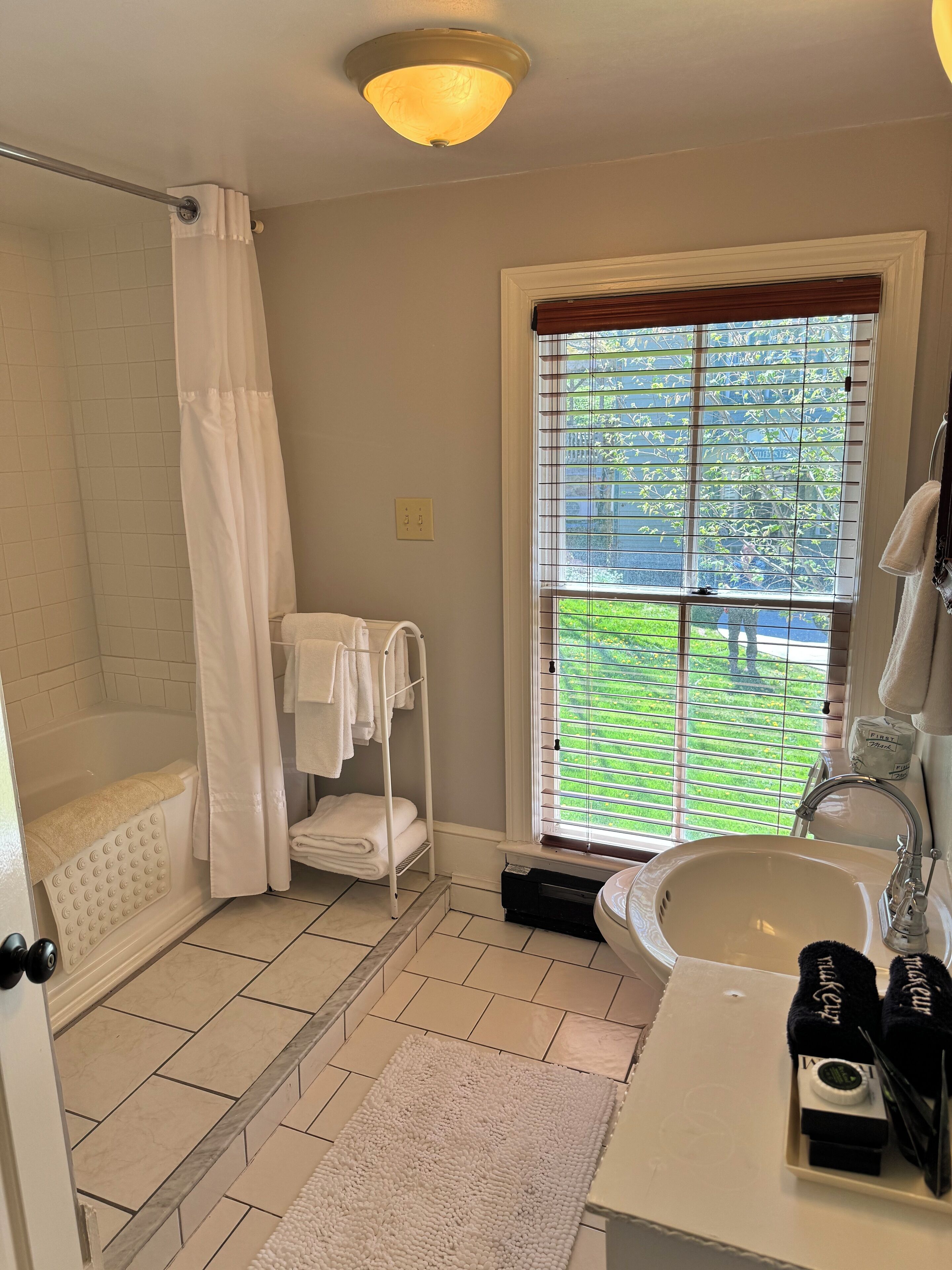 A bathroom with a bathtub, a shower, a window with blinds, a sink, and a towel rack.