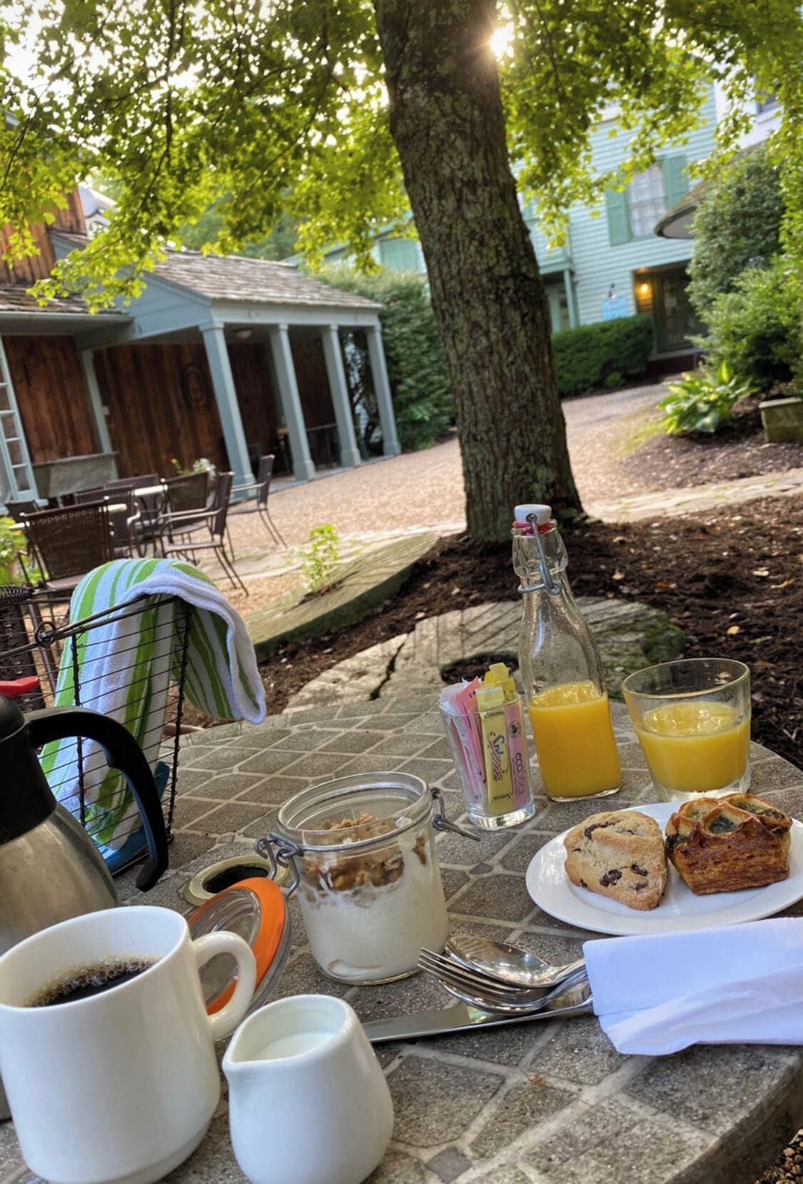 A breakfast table set outdoors with a coffee pot, mugs, juice glasses, a bottle, and pastries.