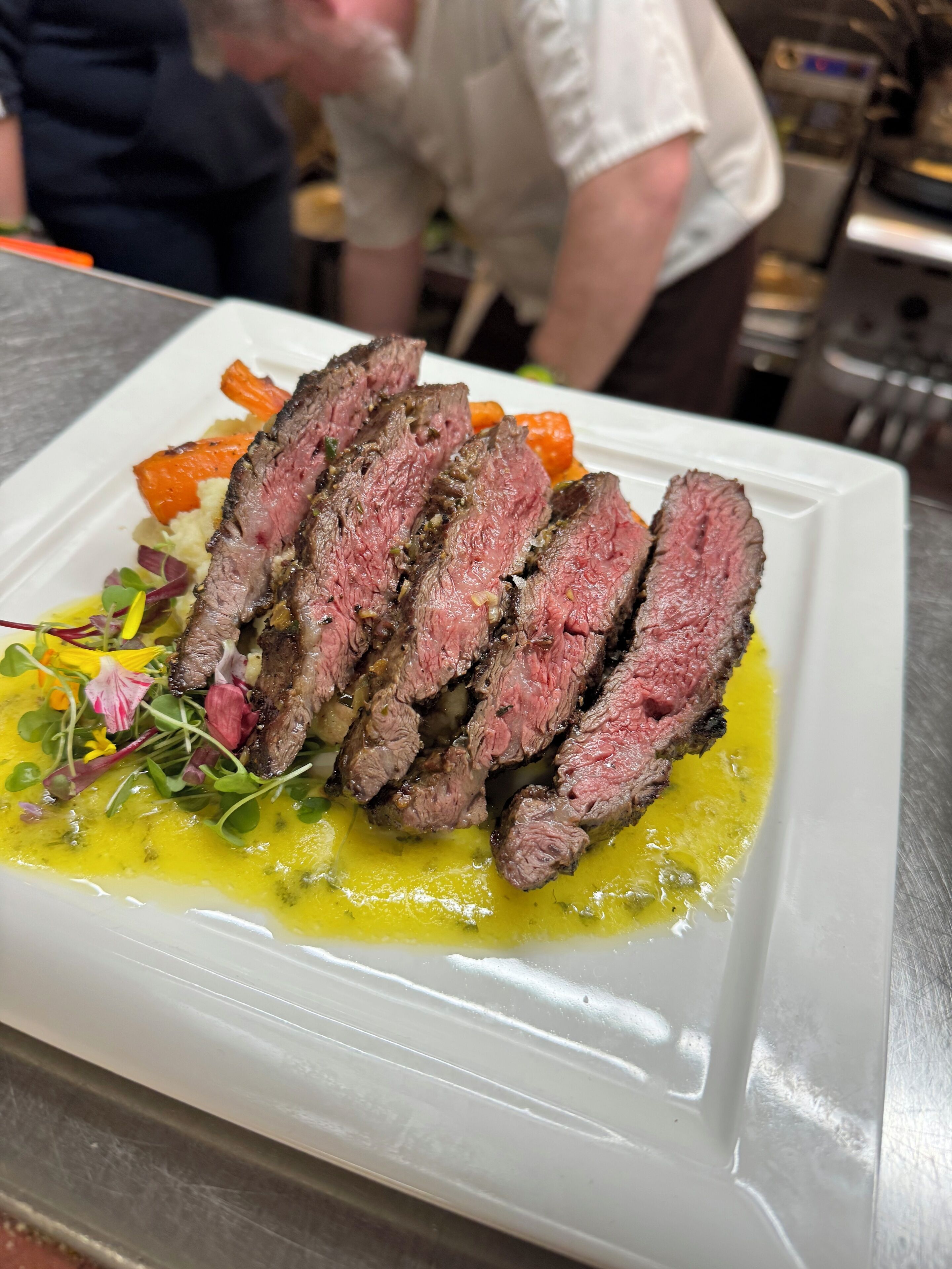 A white rectangular plate with sliced steak, vegetables, and a yellow sauce.