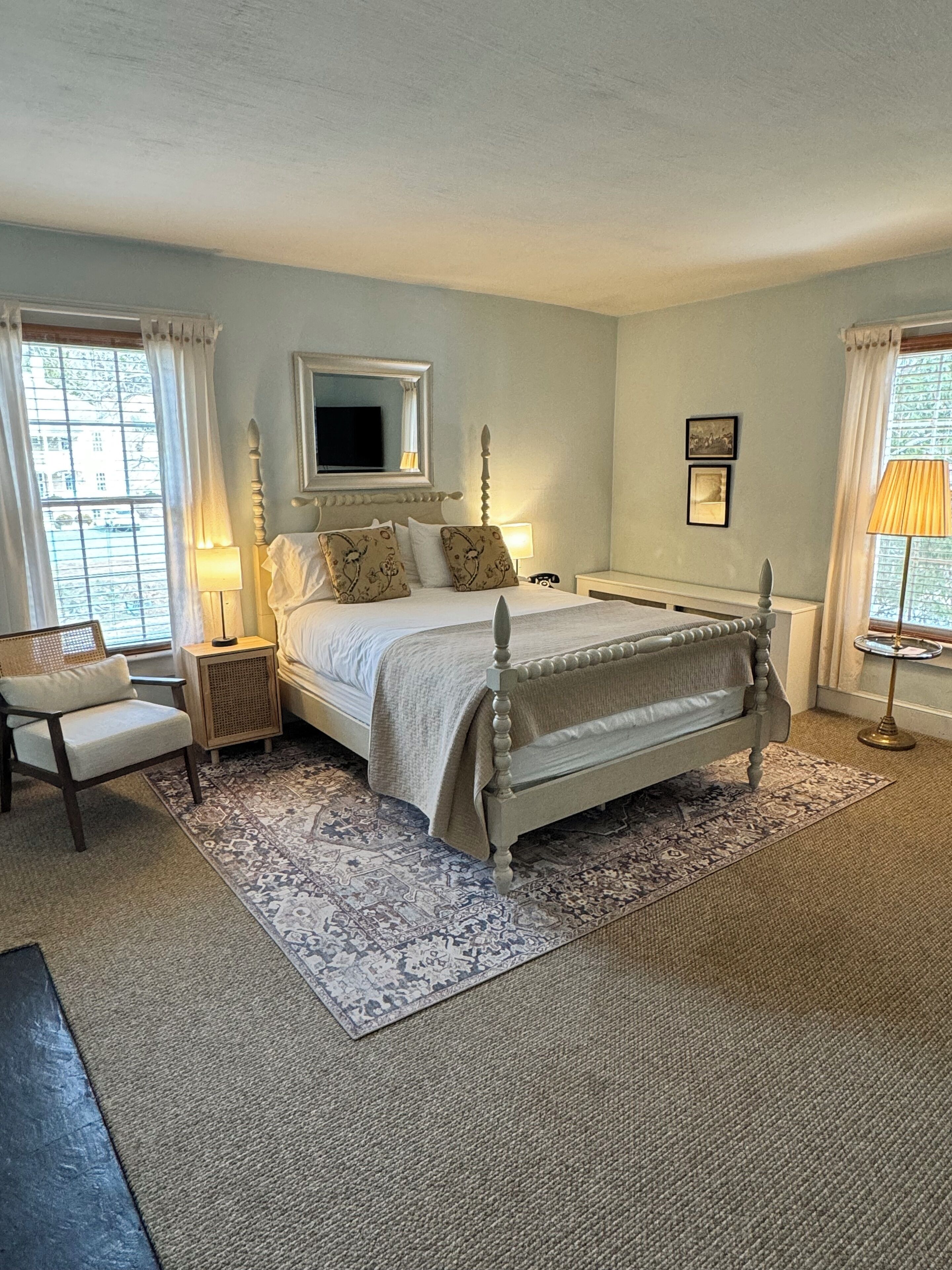 A bedroom with a four-poster bed, a bedside table, a chair, a TV mounted on the wall, and a window with curtains.