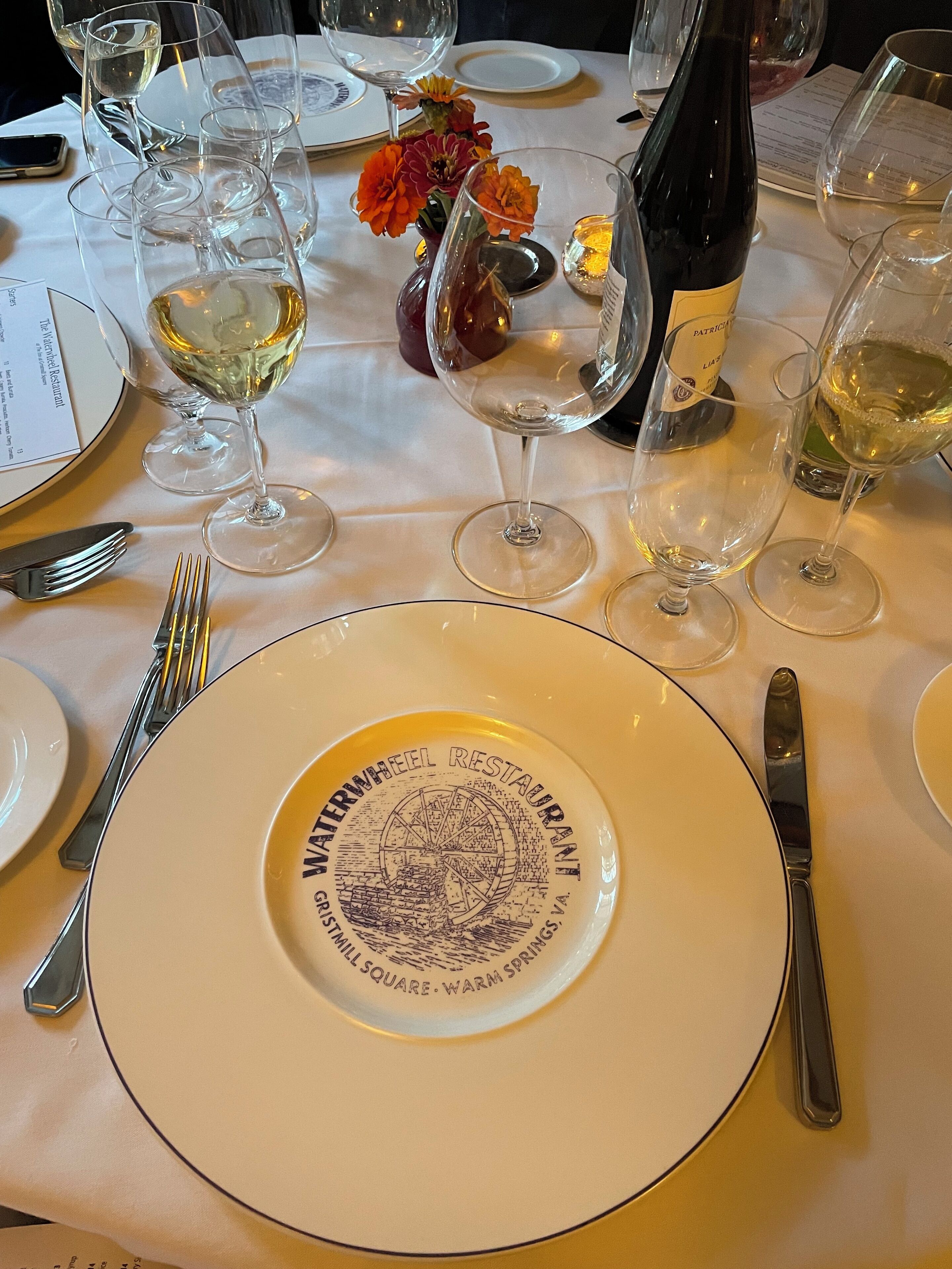 A dining table set with plates, glasses, cutlery, and a bottle of wine.