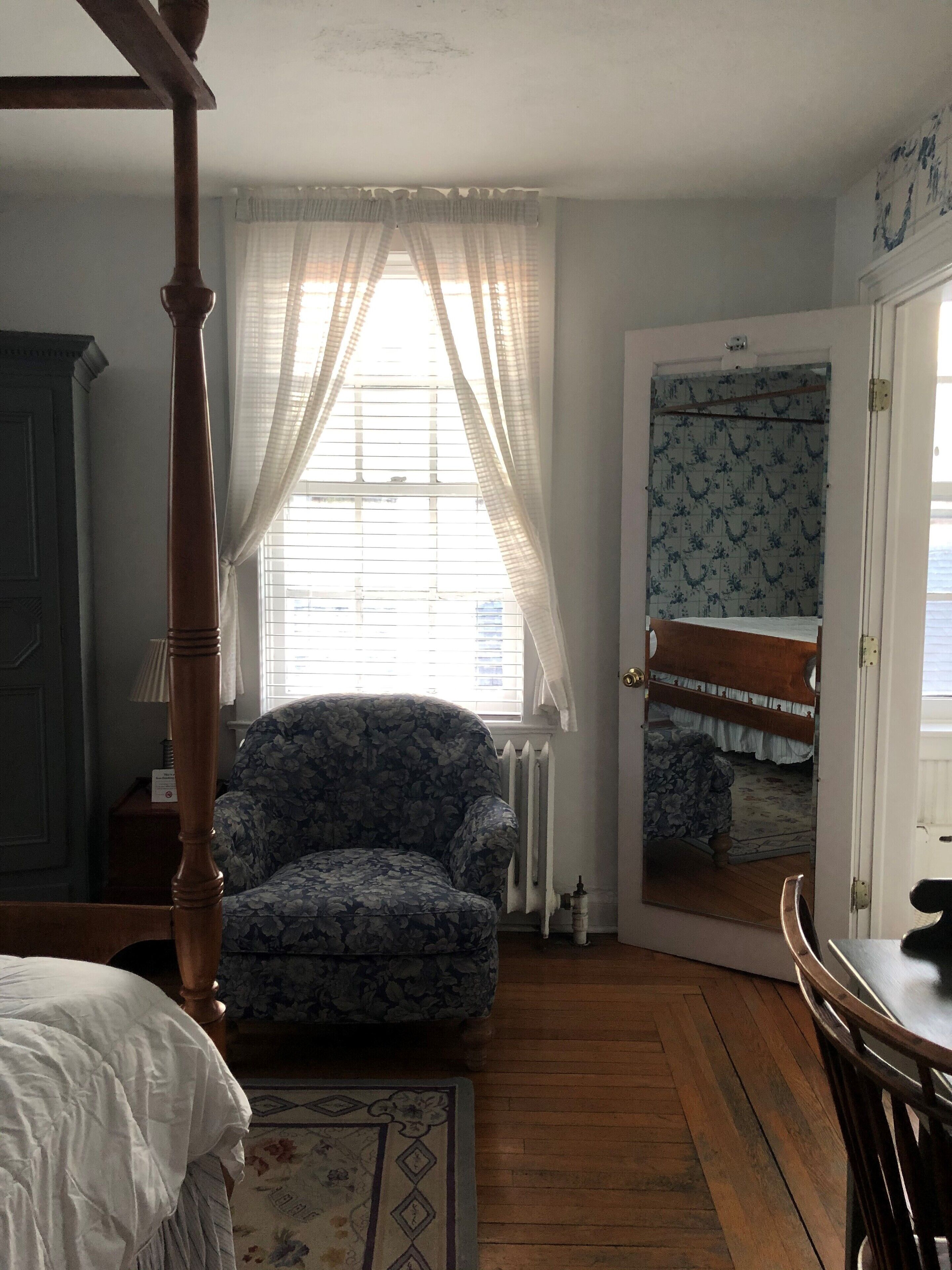 Een slaapkamer met een hemelbed, een fauteuil met bloemenprint, een houten vloer, een radiator en een spiegel op de deur.