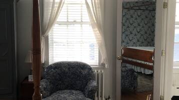 Een slaapkamer met een hemelbed, een fauteuil met bloemenprint, een houten vloer, een radiator en een spiegel op de deur.