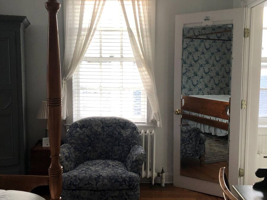 Een slaapkamer met een hemelbed, een fauteuil met bloemenprint, een houten vloer, een radiator en een spiegel op de deur.