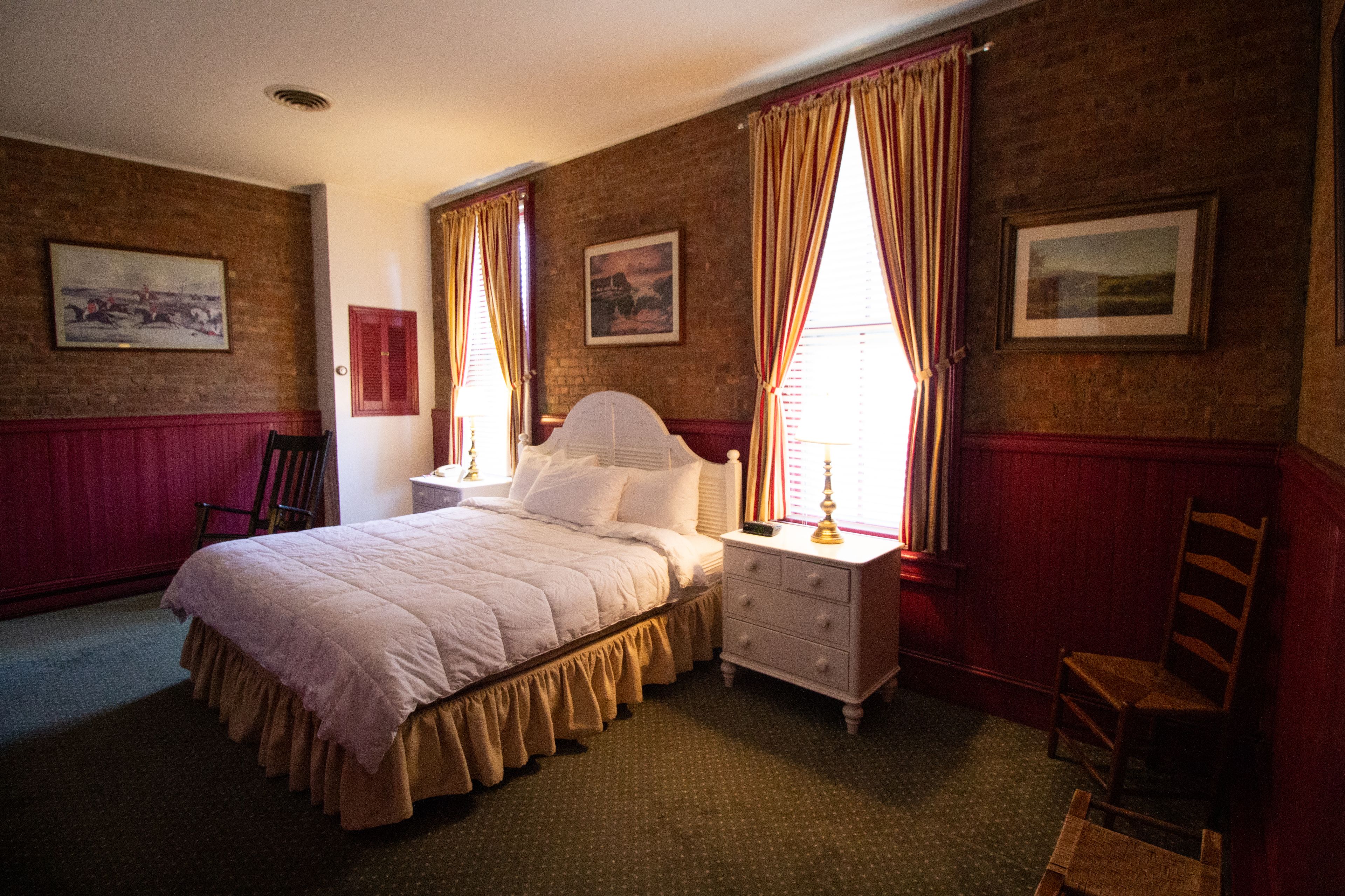 A neatly arranged bedroom with a large bed, a bedside table with a lamp, a chair, and a window with curtains.
