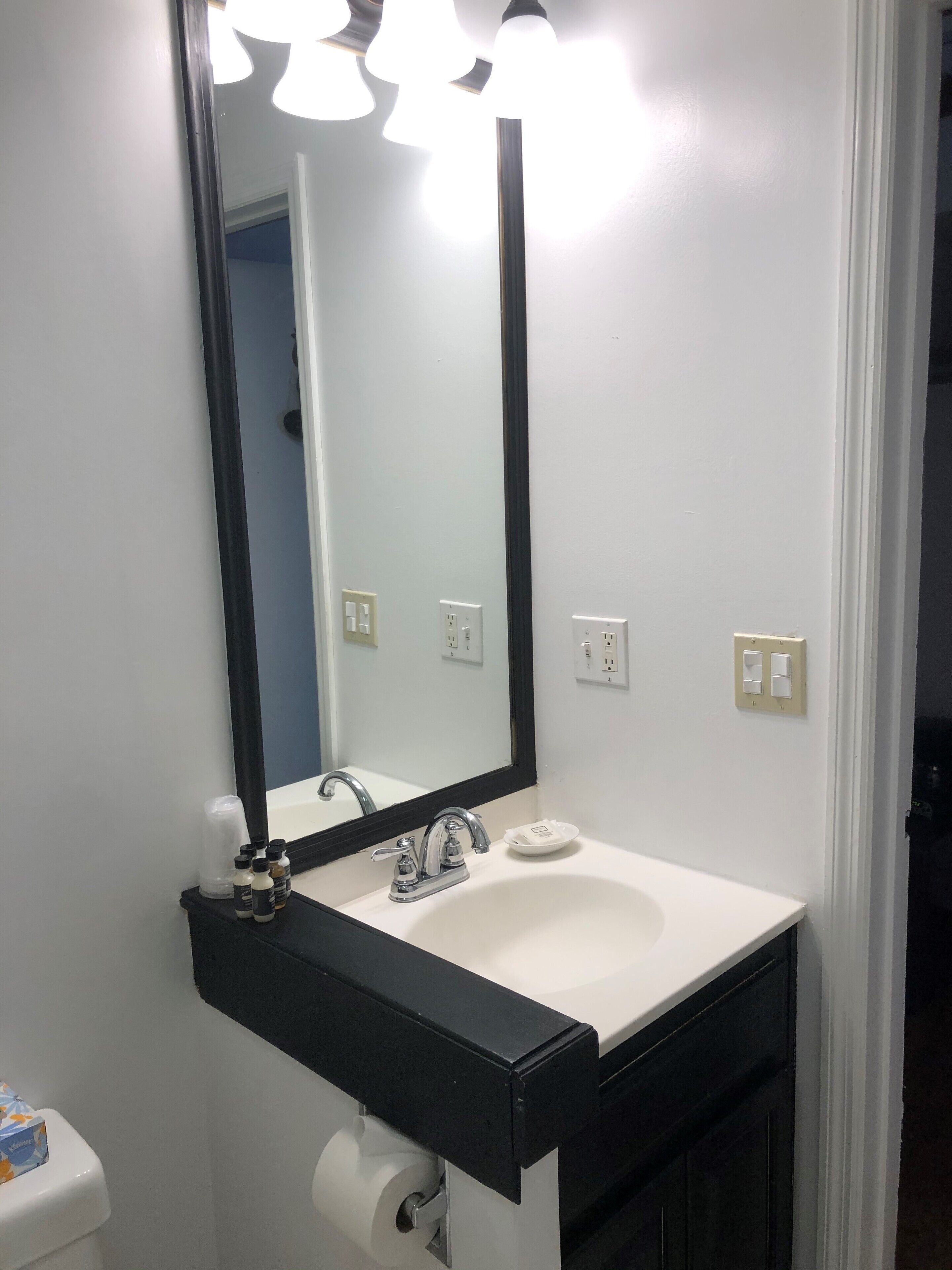 A bathroom with a white sink, black countertop, and a mirror with lights above.