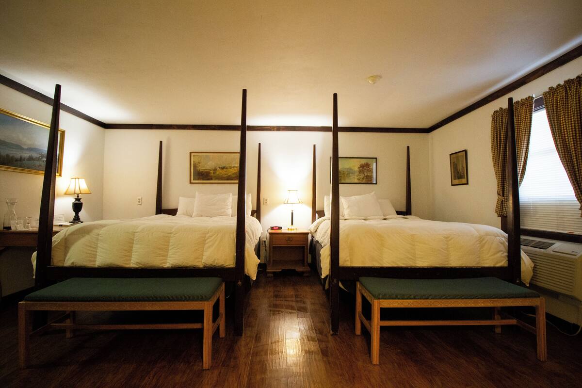 A room with two single beds, a bench, a nightstand, a lamp, and a painting on the wall.