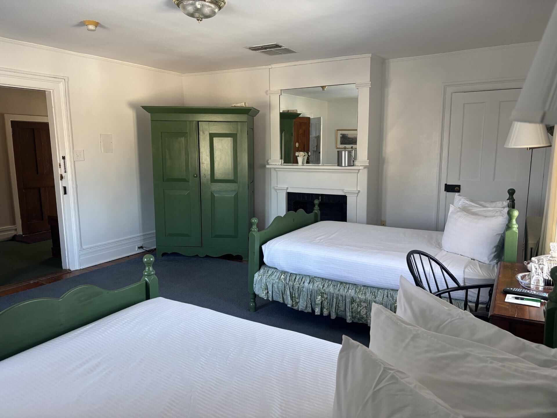 A hotel room with two beds, a green wardrobe, a mirror, a fireplace, and a desk with a lamp.