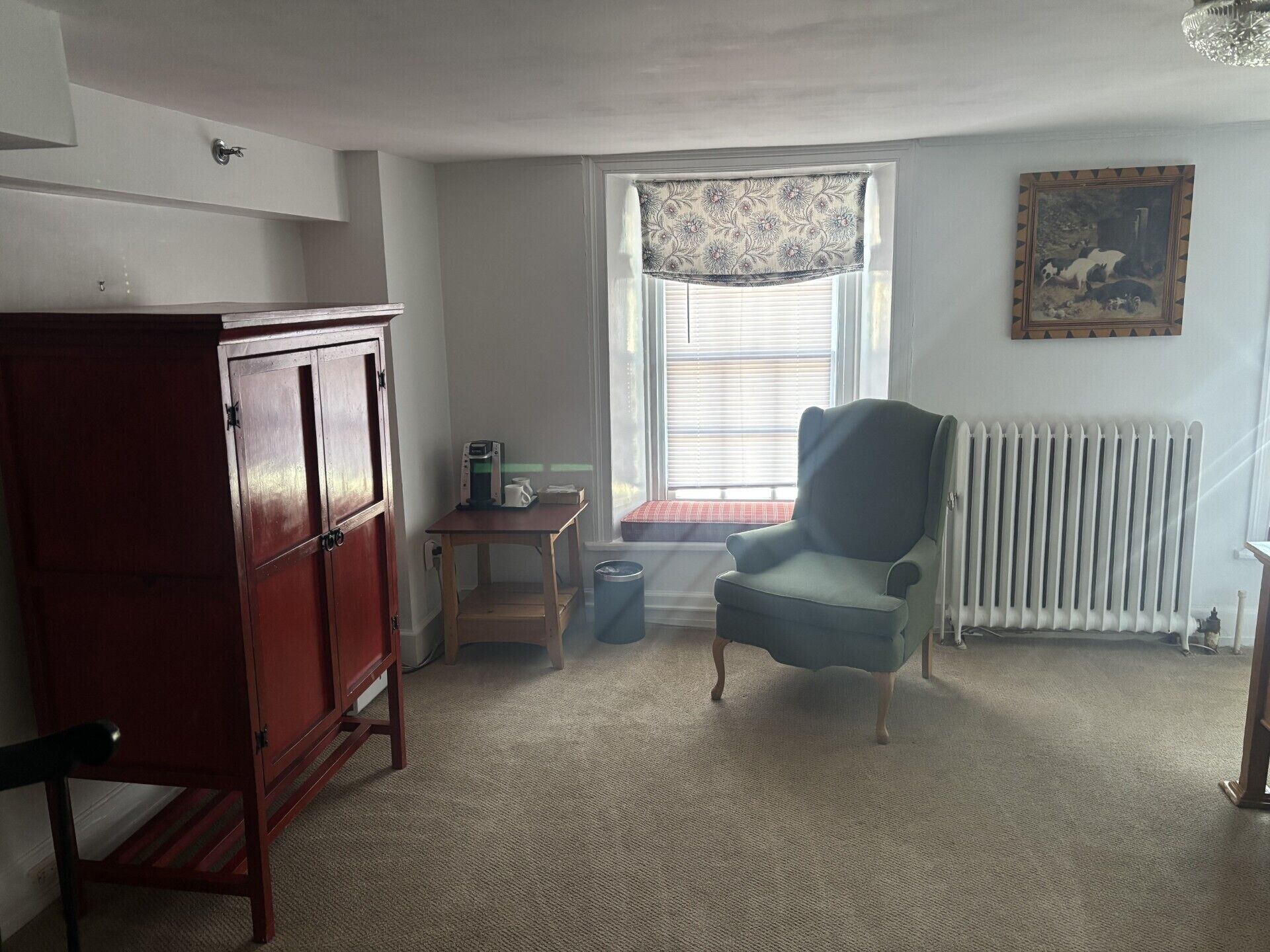 A room with a wooden cabinet, a small table with a coffee maker, a blue armchair, a radiator, and a window with blinds.