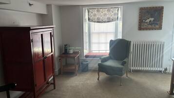 A room with a wooden cabinet, a small table with a coffee maker, a blue armchair, a radiator, and a window with blinds.