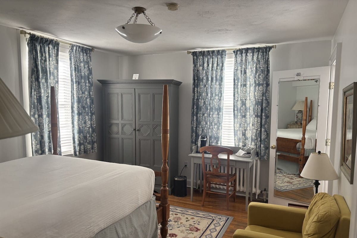 A bedroom with a four-poster bed, a desk with a chair, a yellow armchair, and a large mirror.