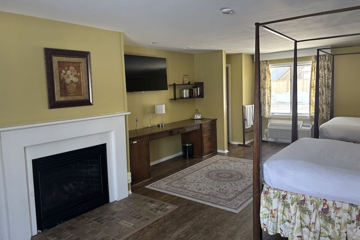A bedroom with a four-poster bed, a fireplace, a desk with a TV, and a window with curtains.