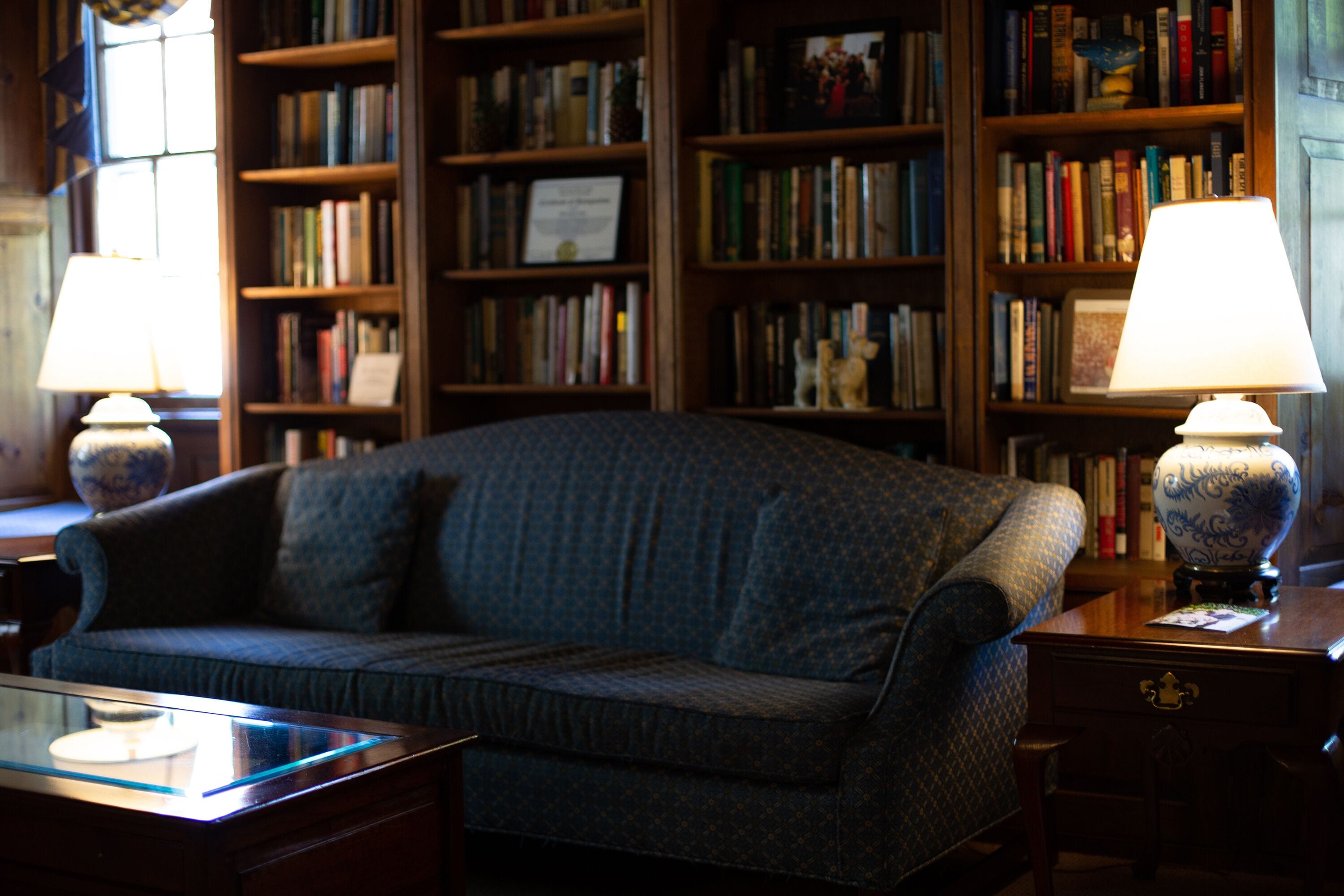 A cozy room with a patterned sofa, a glass coffee table, and two lamps on side tables. A bookshelf filled with books is in the background.