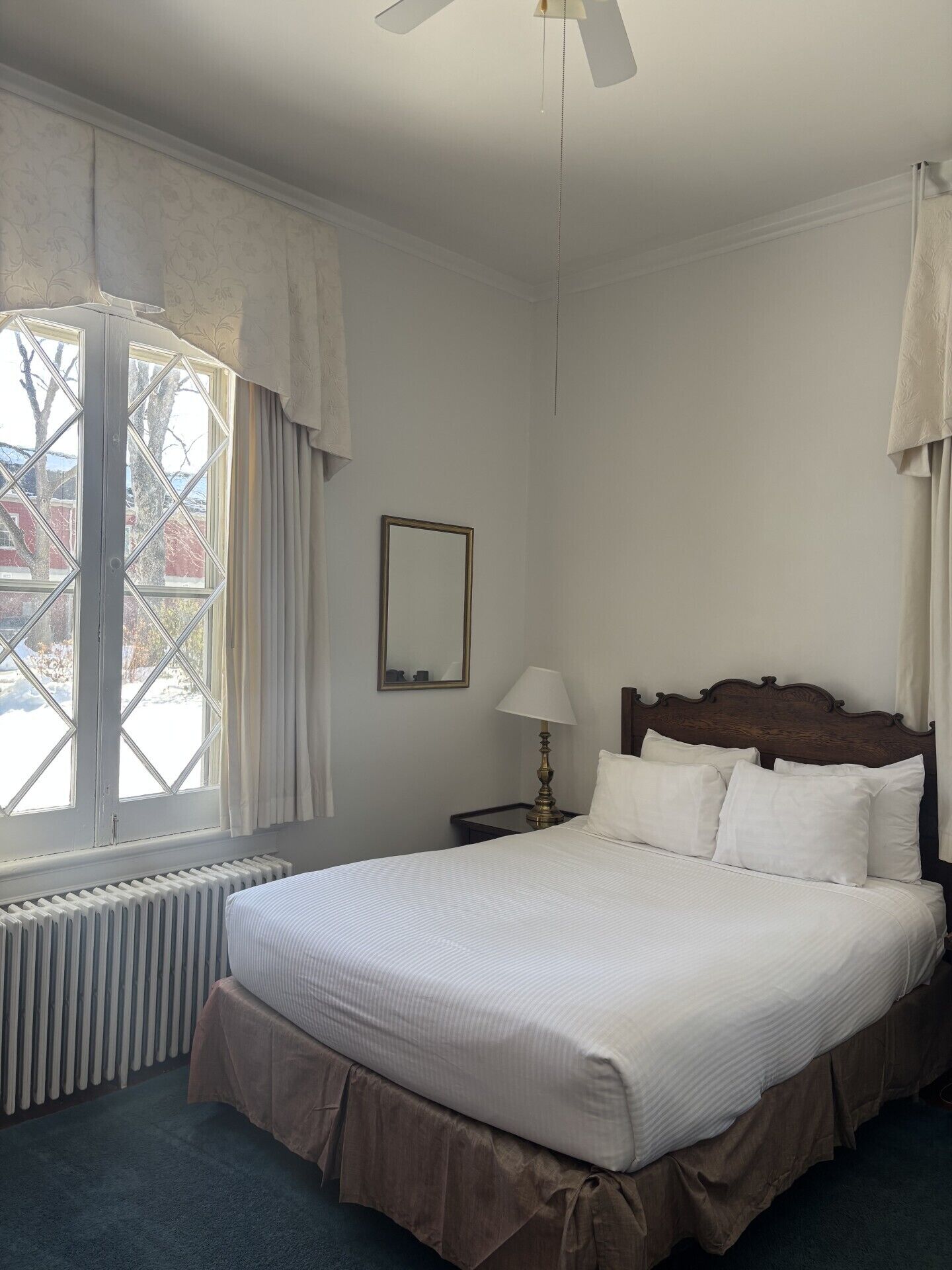 Une chambre d’hôtel avec un grand lit, une table de chevet, une lampe, une fenêtre avec des rideaux et un radiateur.