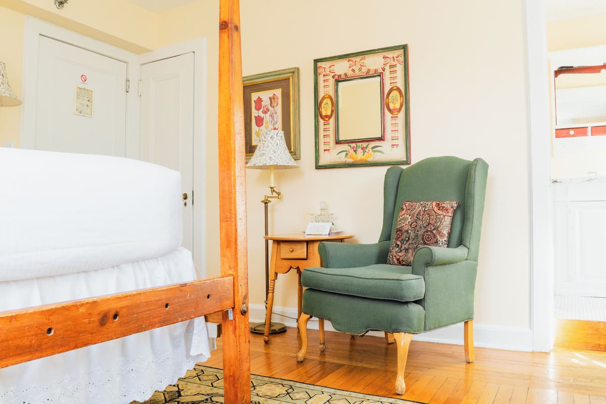 A bedroom with a wooden bed, a green armchair, a small table with a lamp, and two framed pictures on the wall.