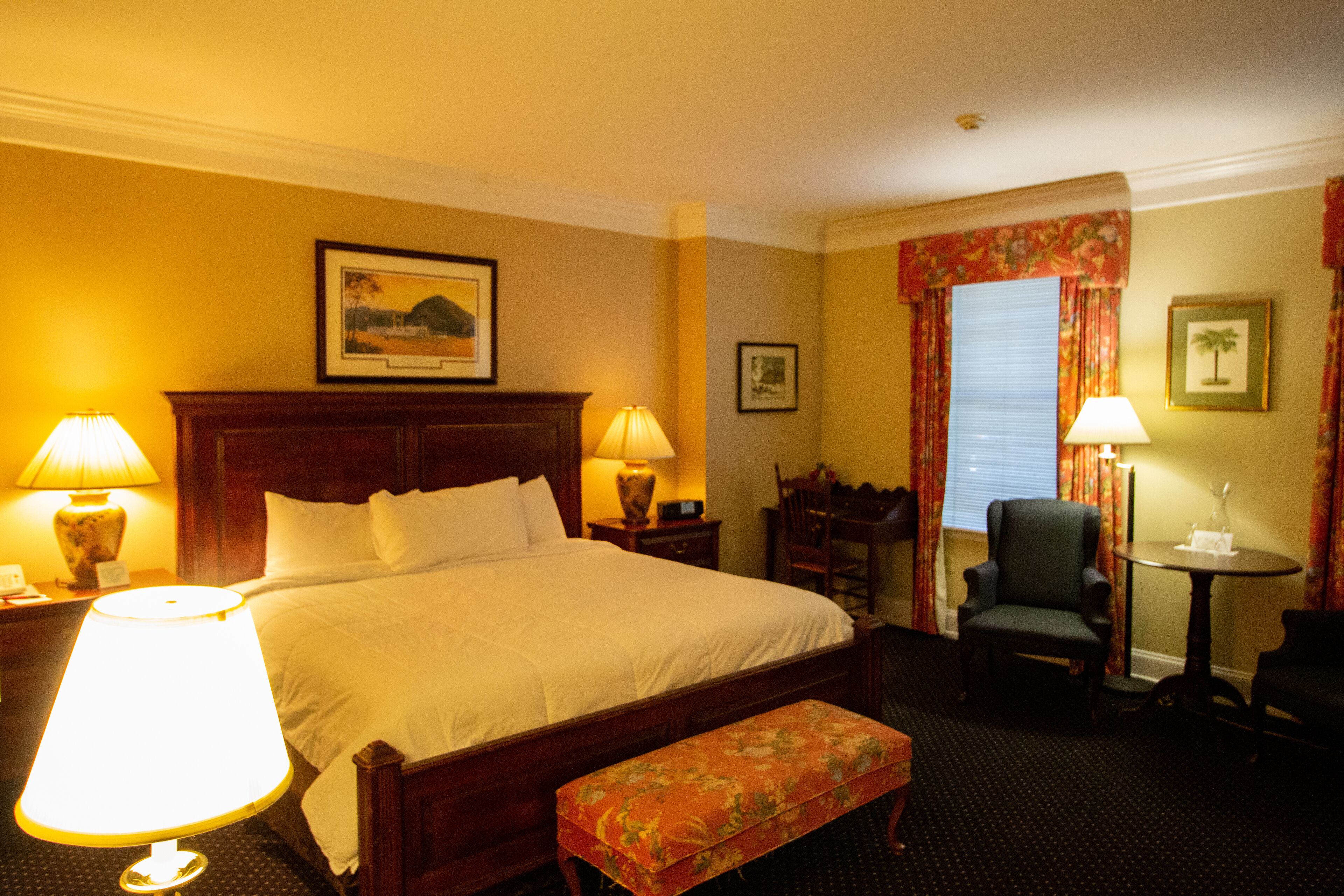 A hotel room with a large bed, two bedside lamps, a wooden headboard, a bench, a chair, a small table, and a window with curtains.