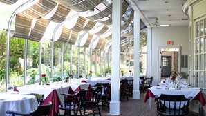 Un espace extérieur abrité, aménagé pour les repas, avec des tables dressées, entouré de colonnes et de grandes fenêtres.