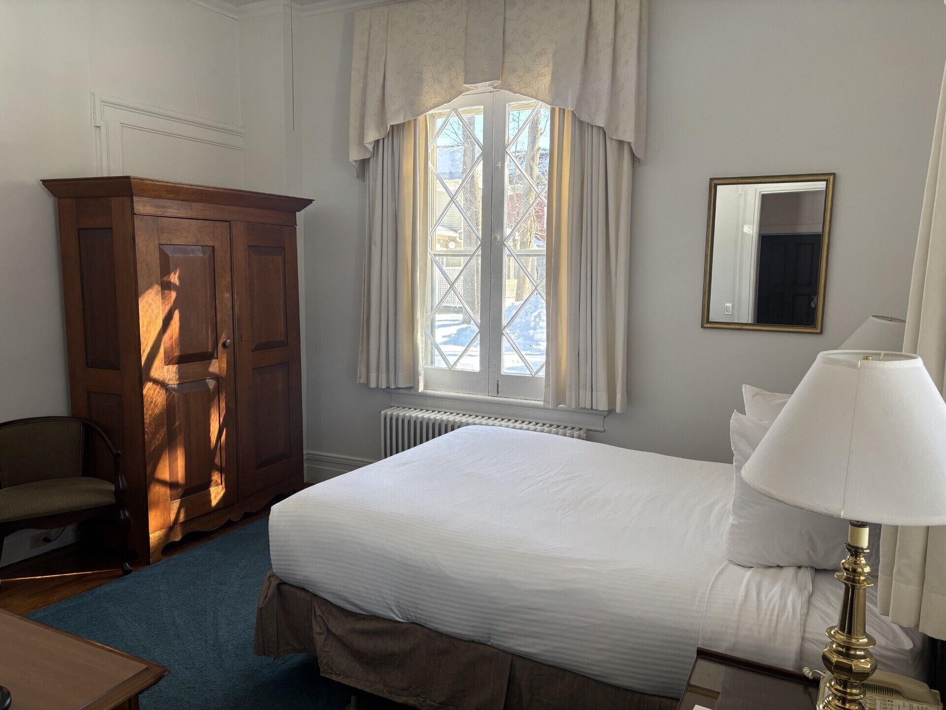A hotel room with a large bed, a wooden wardrobe, a desk, a chair, a window with curtains, and a bedside lamp.