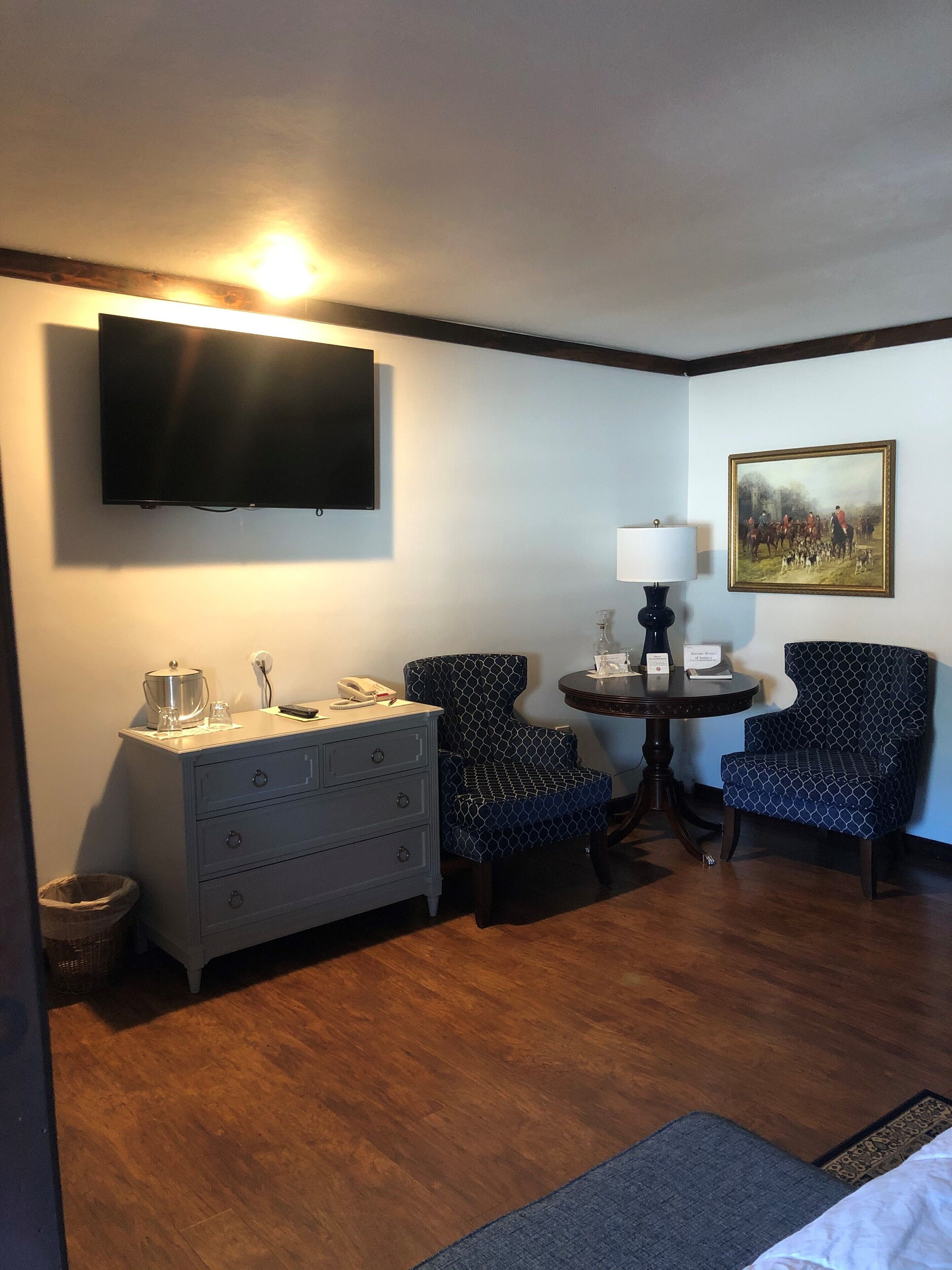 A hotel room with a flat-screen TV, two patterned armchairs, a small round table with a lamp, a dresser, and a painting on the wall.