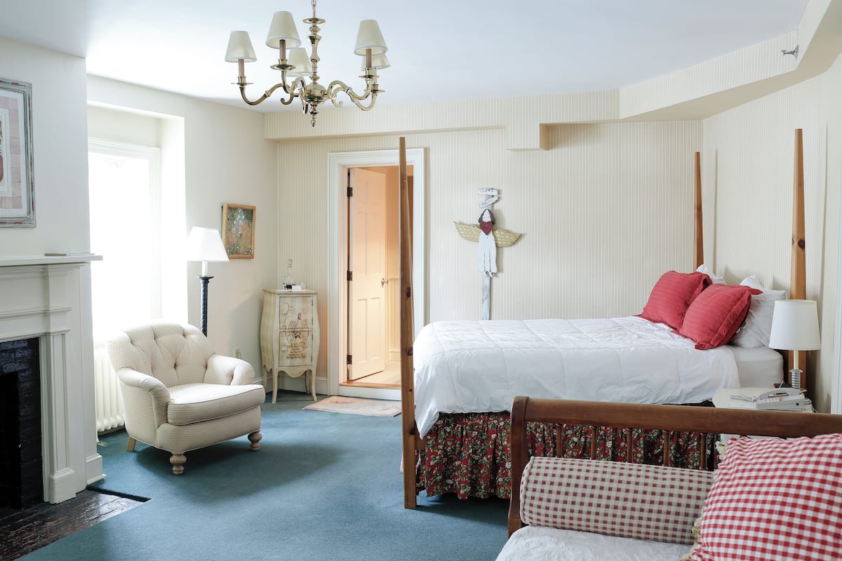 A bedroom with a bed, a chair, a nightstand, a lamp, a fireplace, and a door leading to another room.