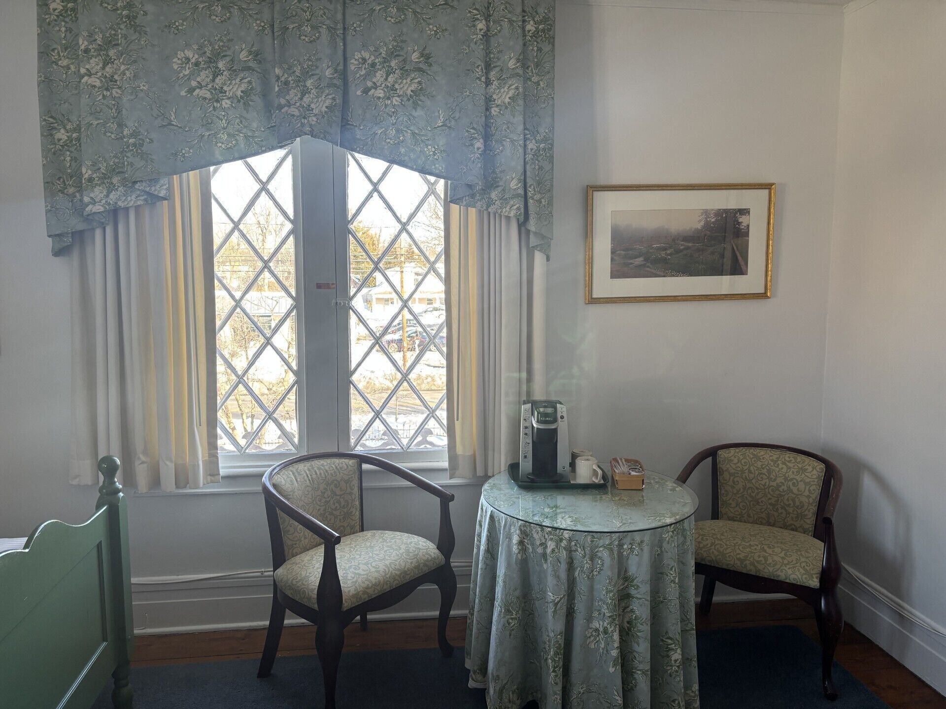 A room with a large window, two chairs, a small table with a coffee maker, and a framed picture on the wall.