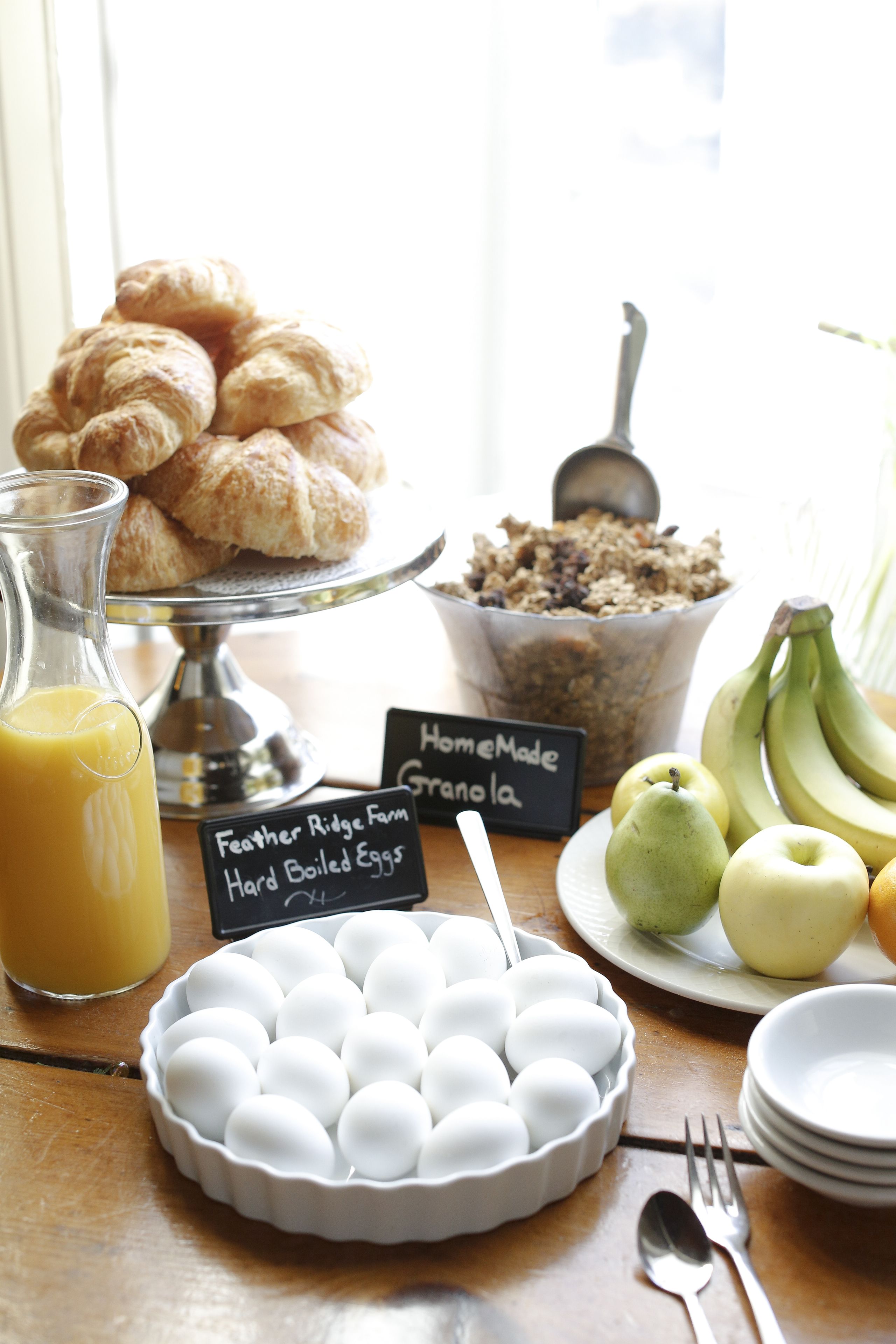 A breakfast table with croissants, a bowl of granola, a pitcher of orange juice, a plate of eggs, and fresh fruit.