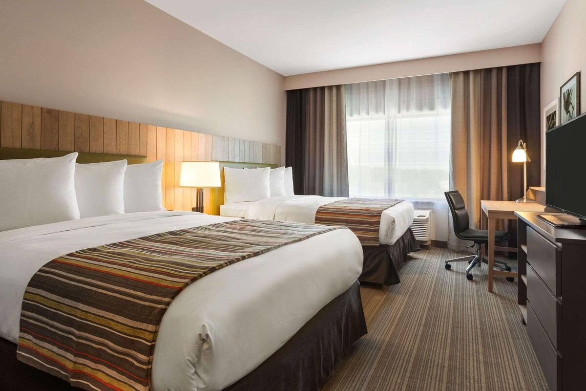 Ein Hotelzimmer mit zwei Betten, einem Schreibtisch, einem Stuhl, einer Lampe und einem großen Fenster mit Vorhängen.