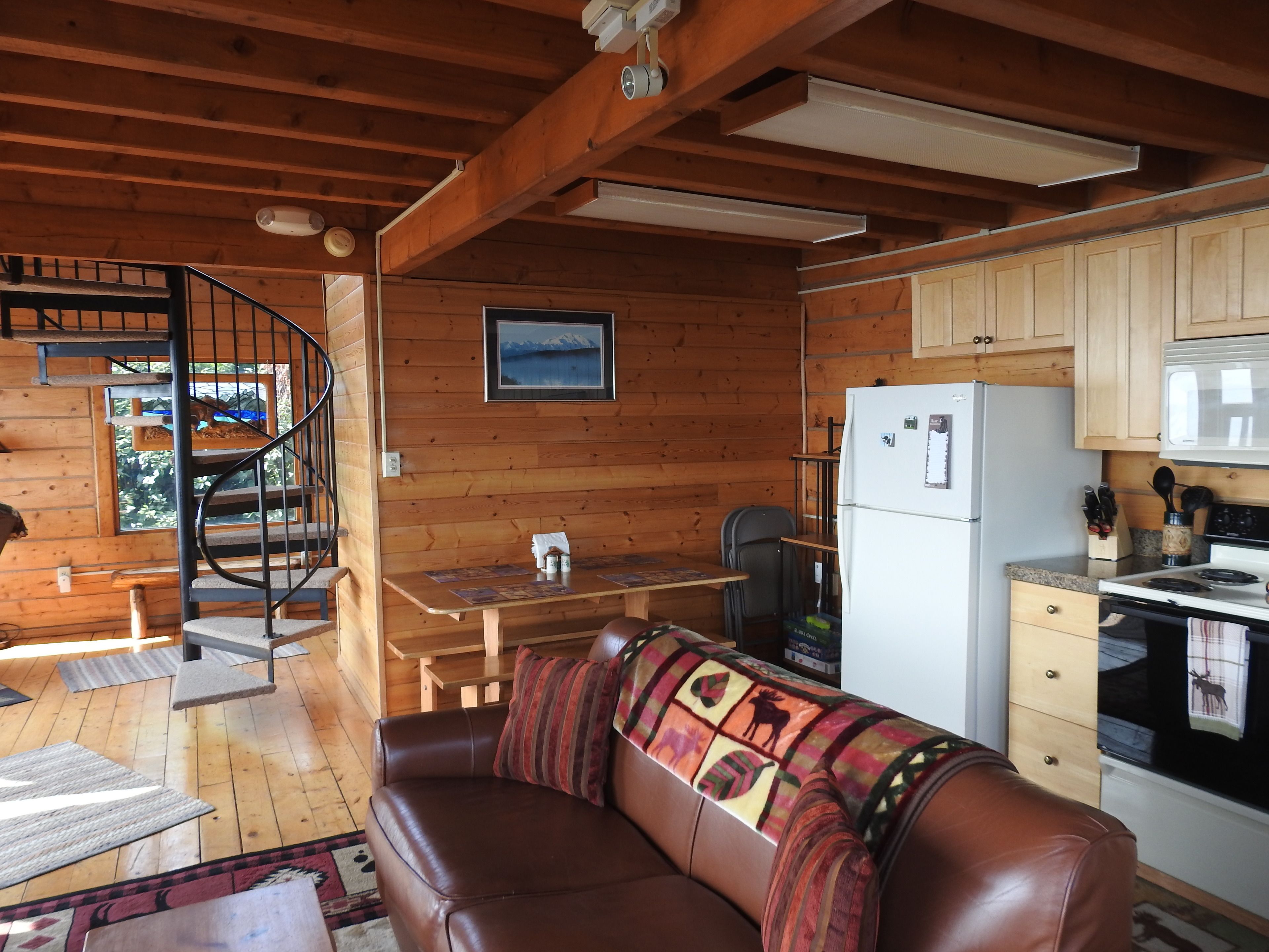 A cozy cabin interior with a wooden interior, a sofa, a dining table, a kitchen area, and a spiral staircase.