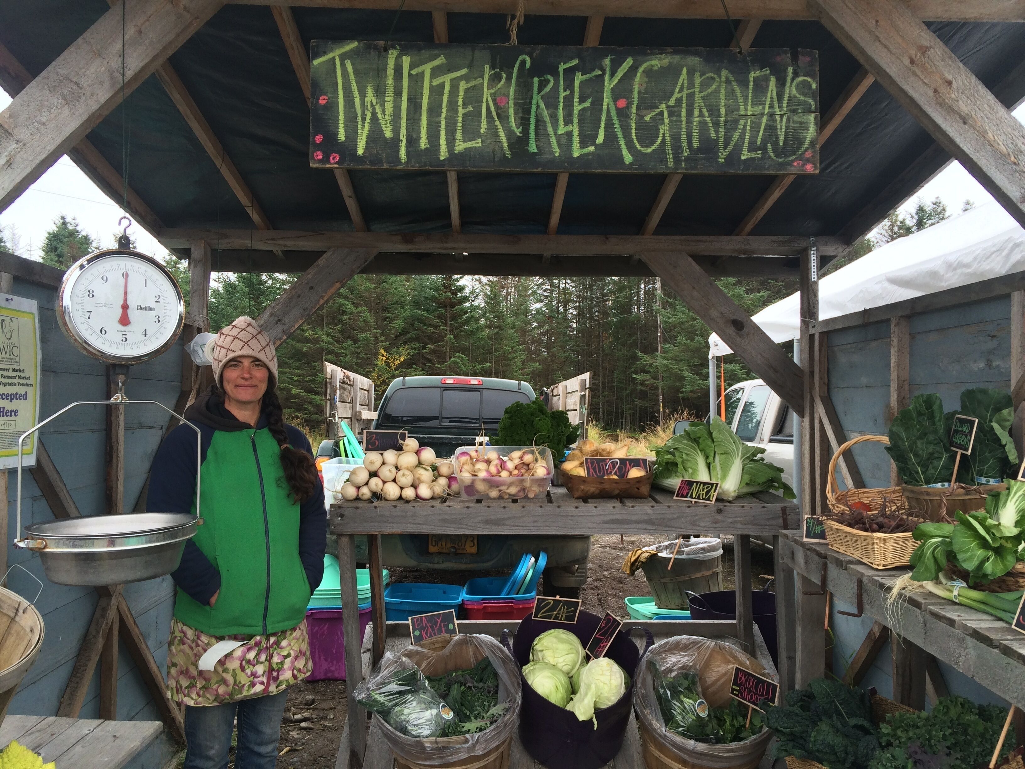 Un étal de marché avec une pancarte indiquant « JARDINS DE TWITTER CREEK » et diverses sortes de légumes exposées.