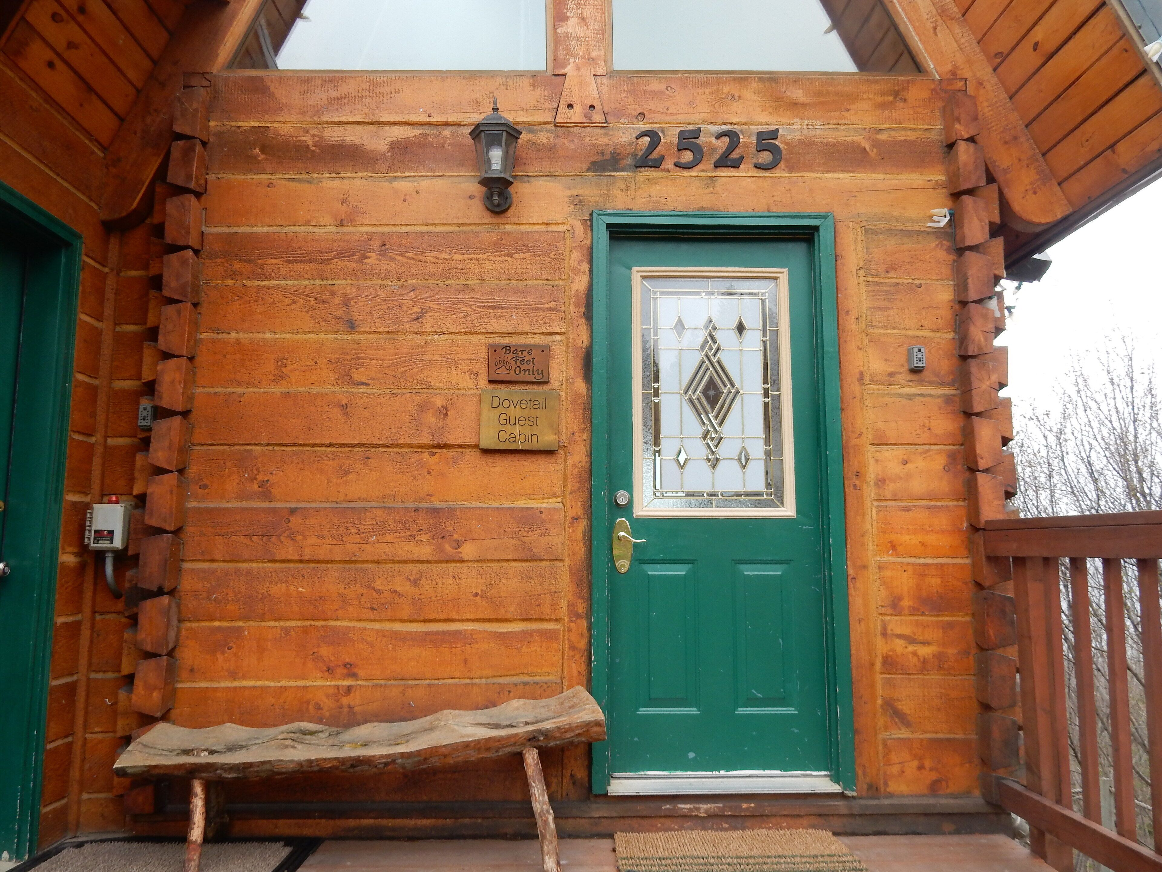 Una cabaña de madera con puerta verde, un banco afuera y un letrero que indica que es la Cabaña de Huéspedes Dovetail.