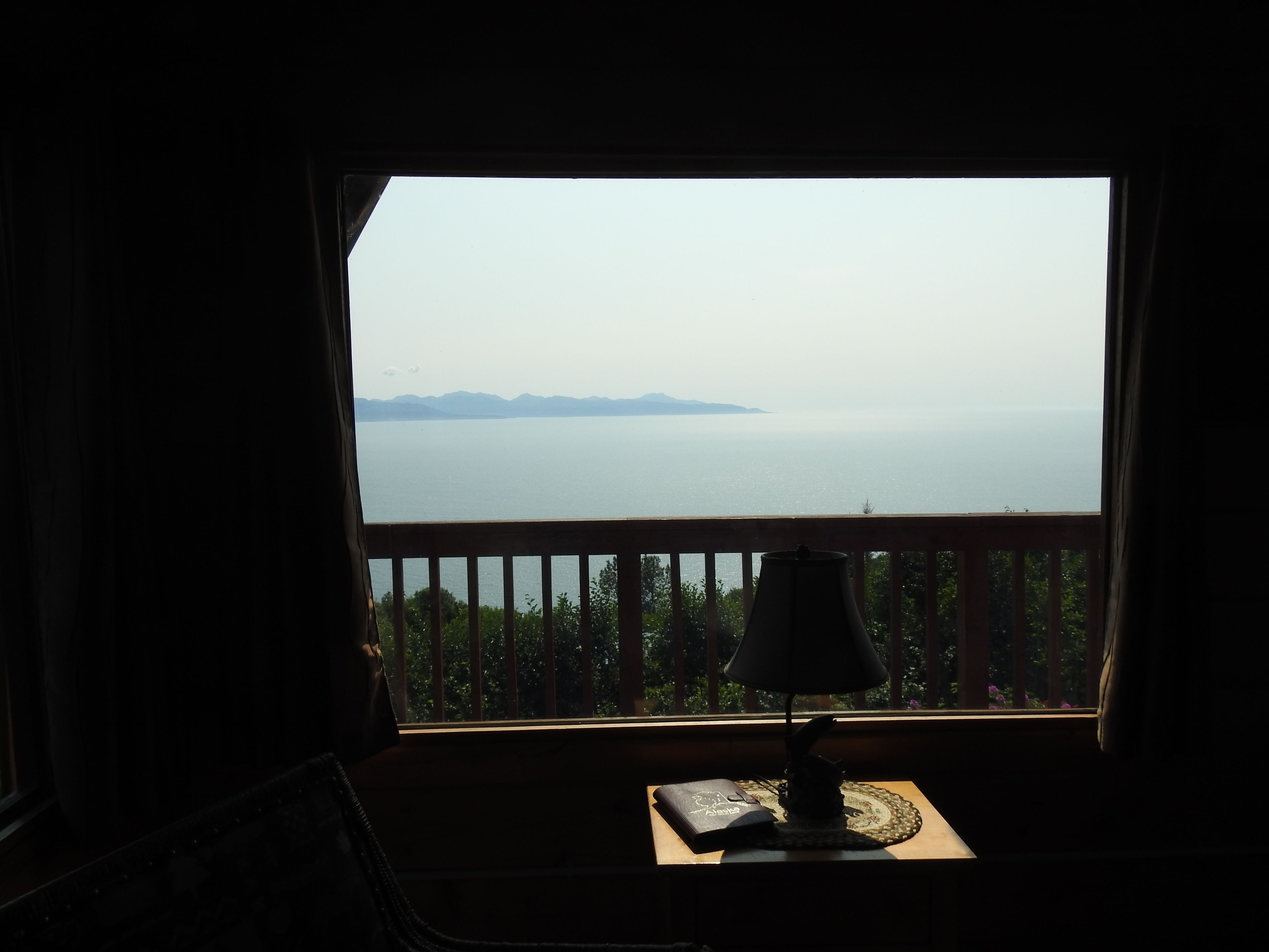 Ein Zimmer mit Blick auf ein großes Gewässer und ferne Berge. Es gibt ein Holzgeländer und einen Tisch mit einer Lampe und einem Buch.