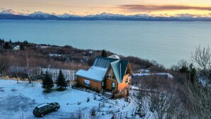 Une cabane au toit en pente, entourée de neige, située près d’une étendue d’eau, avec des montagnes en arrière-plan.