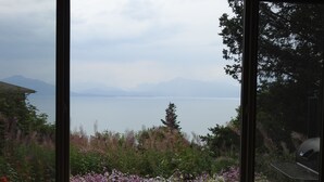 Une vue à travers une fenêtre sur un lac, avec des montagnes en arrière-plan, le tout entouré d’une végétation luxuriante et de fleurs.