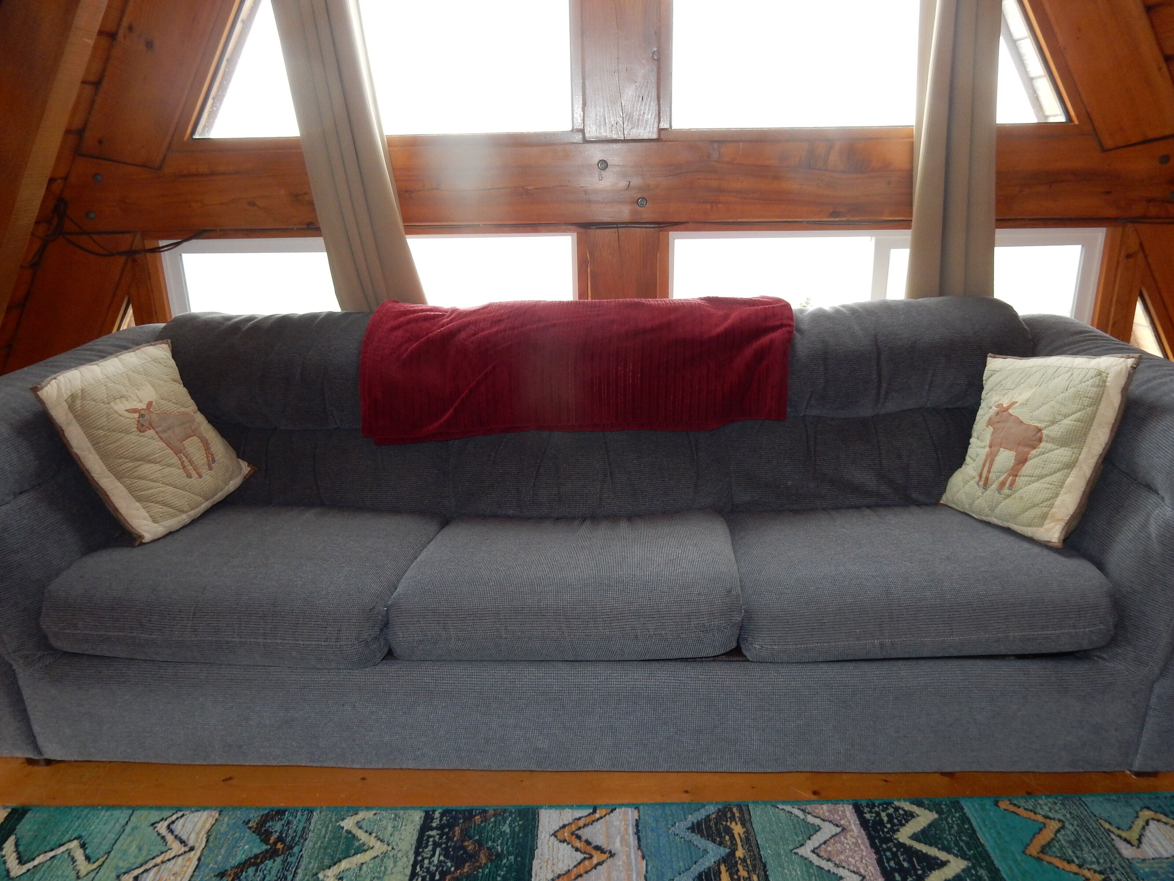 A grey sofa with two decorative pillows, a red blanket, and a patterned rug in a room with wooden beams and large windows.