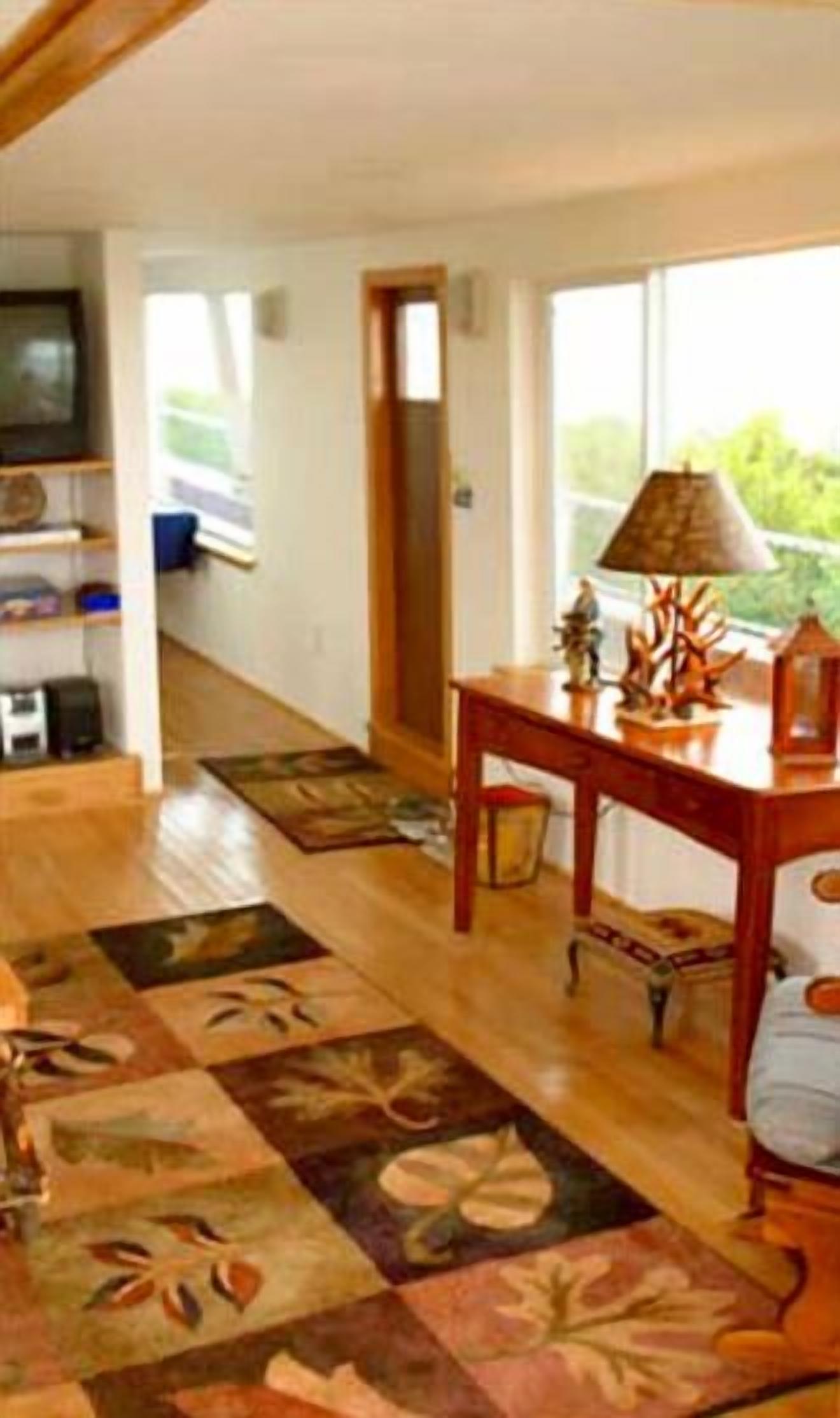 A living room with a wooden floor, a television, a table with decorative items, and a view of the outdoors through large windows.
