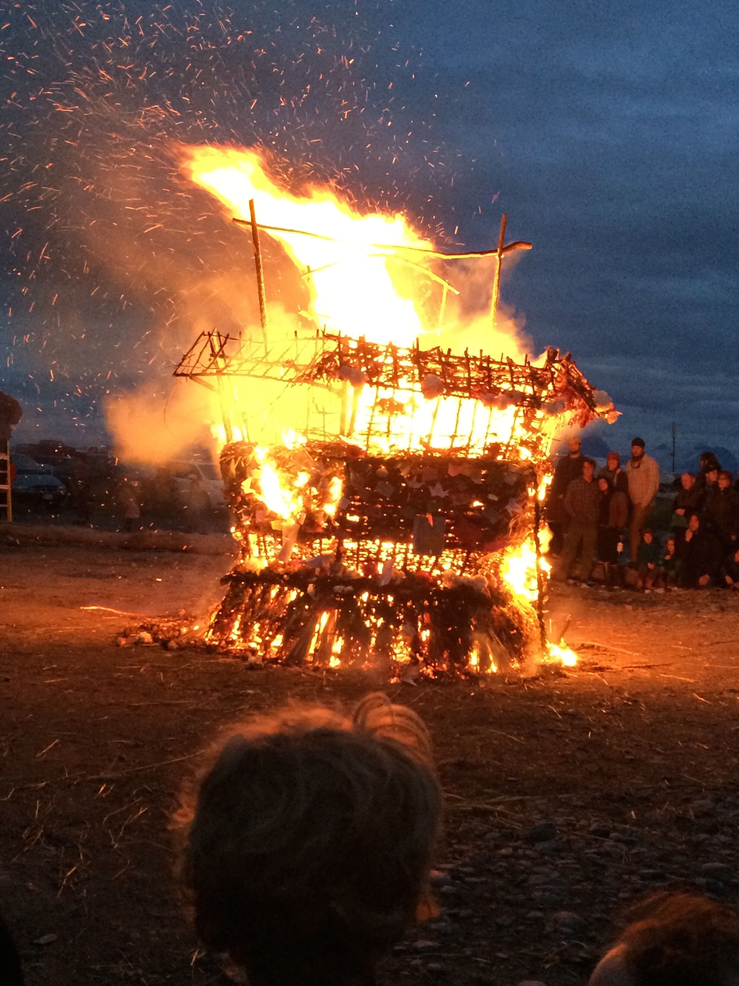 Un grand feu de joie surmonté d’une structure métallique, autour duquel se rassemblent des personnes.