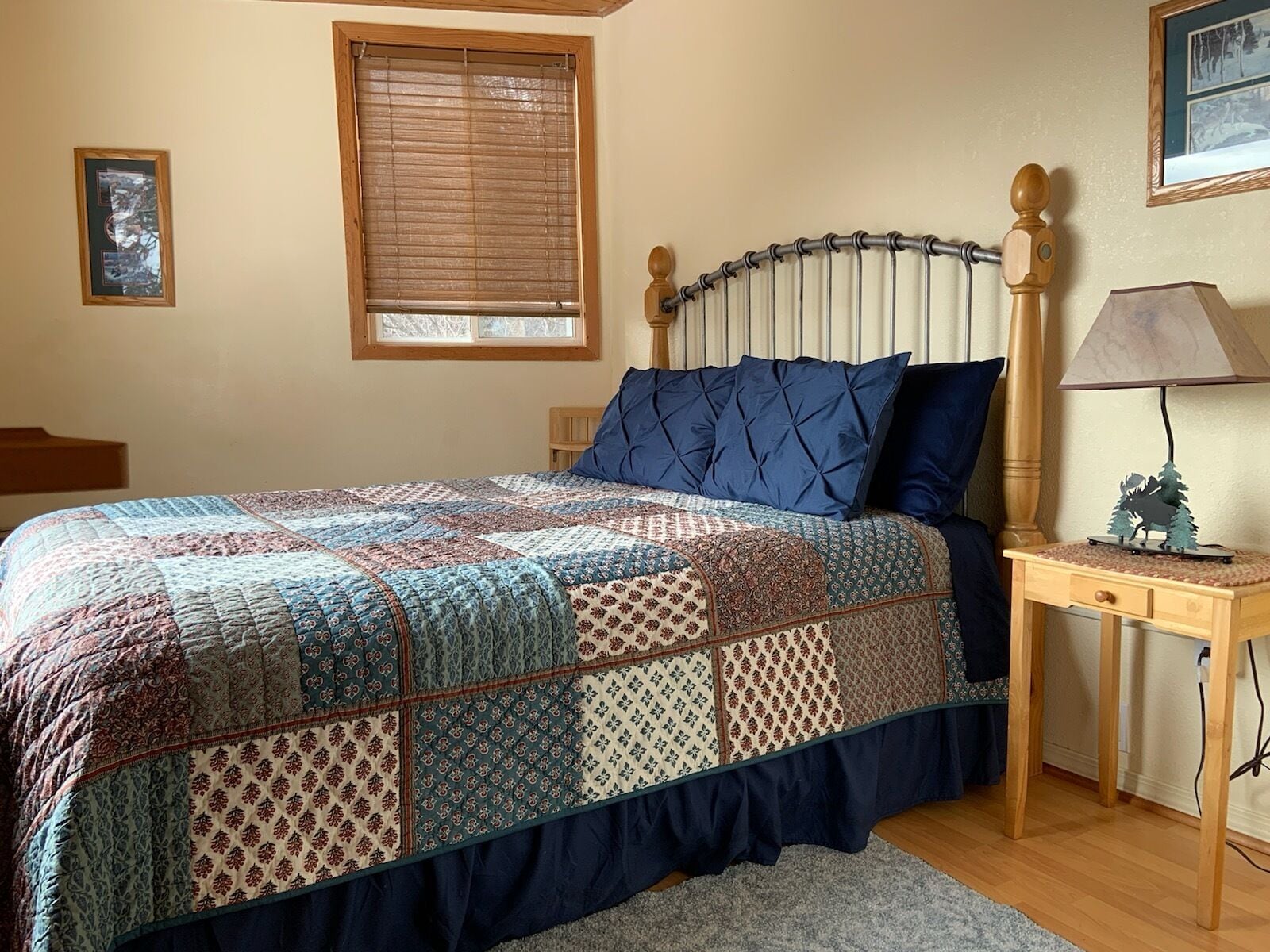 A bedroom with a bed, a nightstand, a lamp, a window with blinds, and a framed picture on the wall.