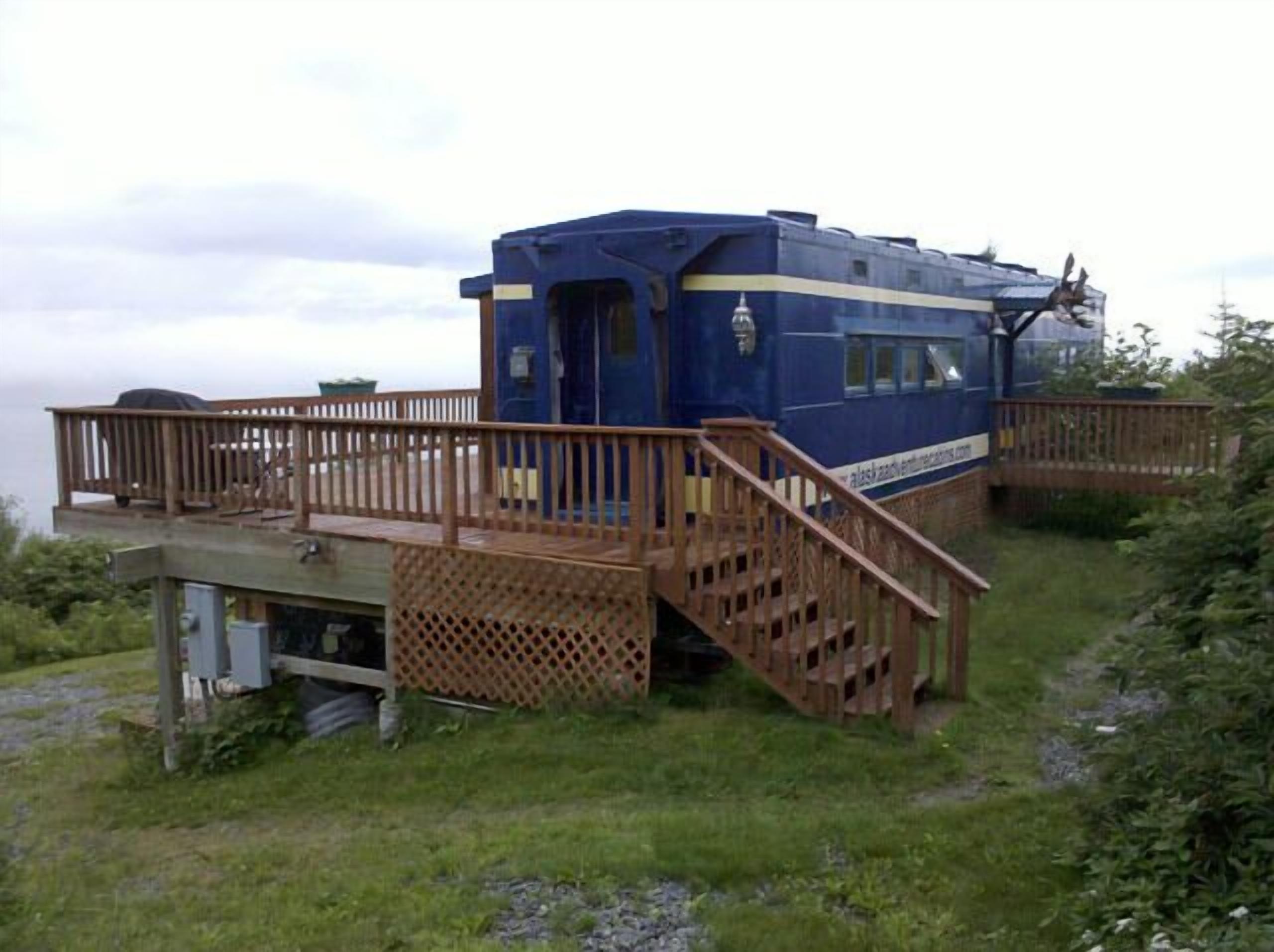 A blue train car converted into a cabin with a wooden deck and stairs leading to the entrance.