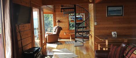 Ein gemütliches Blockhäuschen-Interieur mit Holzausbau, Kamin, einer Couch, einem Esstisch und einer Wendeltreppe.